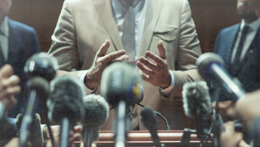 Header-Bild mit Oberkörper eines Mannes im Anzug, vor ihm zahlreiche Presse-Mikrofone, die auf ihn gerichtet sind.