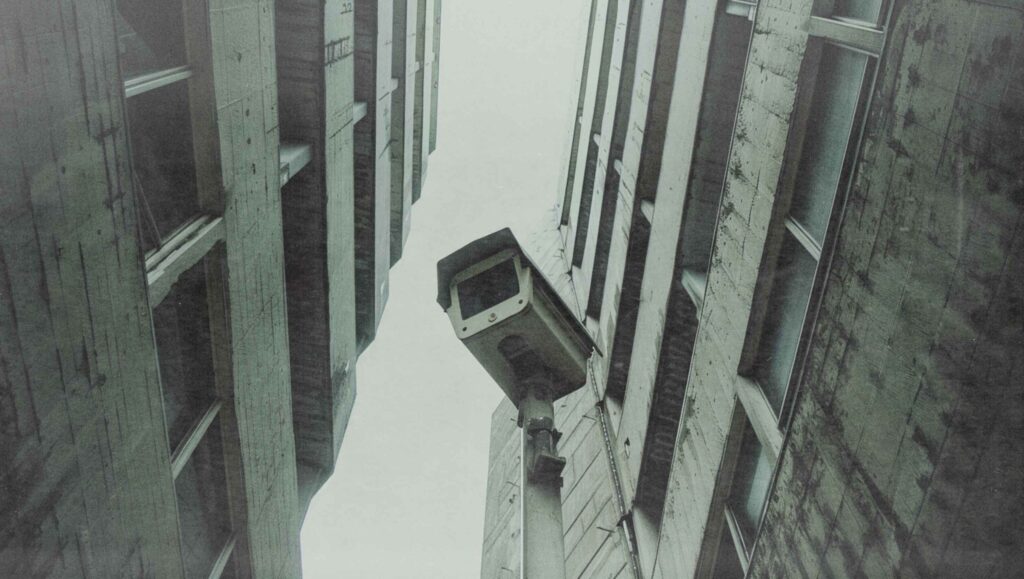 Videokamera im öffentlichen Raum, von unten fotografiert zwischen zwei Betonhochhäusern mit Blick in den Himmel.