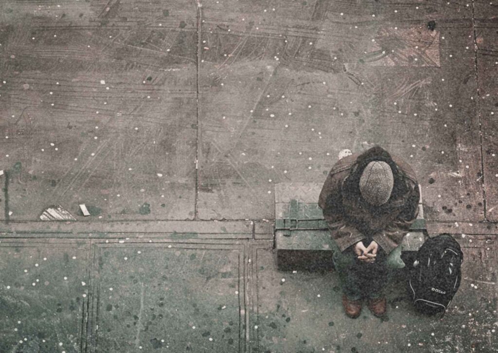 Von oben aufgenommenes Foto: Ein Mann sitzt rechts im Bild mit gesenktem Kopf und gefalteten Händen auf einer Bank. Der Rest des Bildes zeigt nur leeren Beton. Symbolfoto für Armut, Einsamkeit und Hoffnungslosigkeit.
