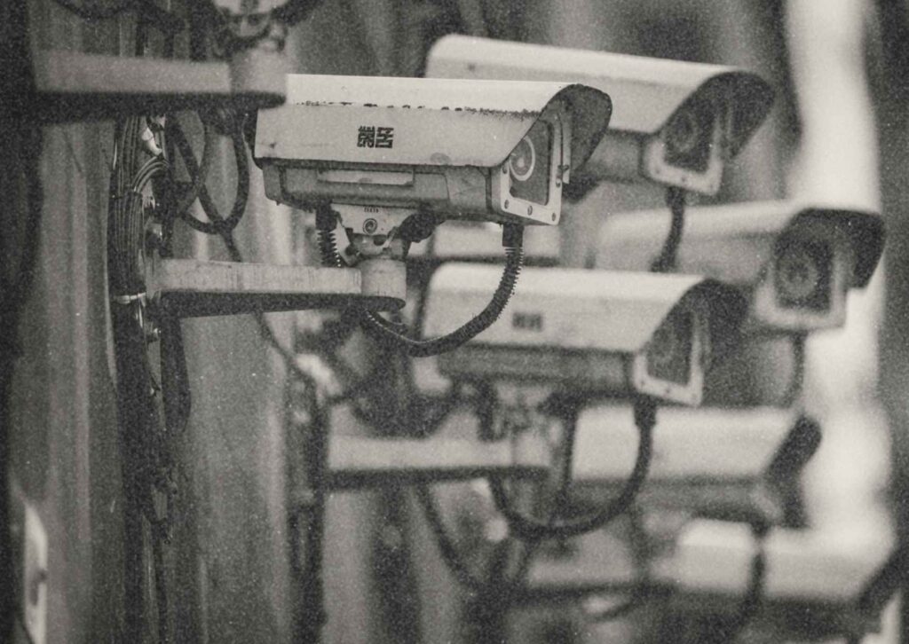 Foto von mindestens sechs Überwachungskameras, die an einer Gebäudefassade im öffentlichen Raum montiert sind.