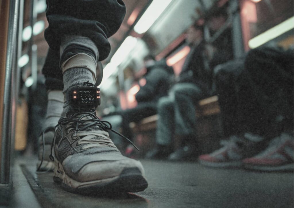 Nahaufnahme eines Fußes mit elektronischer Fußfessel in einer U-Bahn. Im Hintergrund sitzen Menschen unscharf auf den Sitzen.