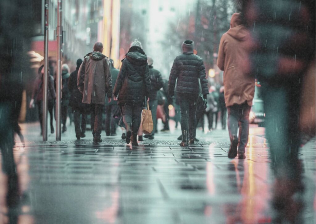 Menschen gehen an einem regnerischen Tag durch eine Stadt. Das Bild zeigt den gesellschaftlichen Alltag als Ausgangspunkt sozialer Dynamiken und präventiver Überlegungen.