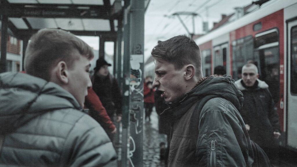 Zwei Jugendliche in angespannter Konfrontation an einer Bahnhaltestelle, im Hintergrund ein einfahrender Zug.